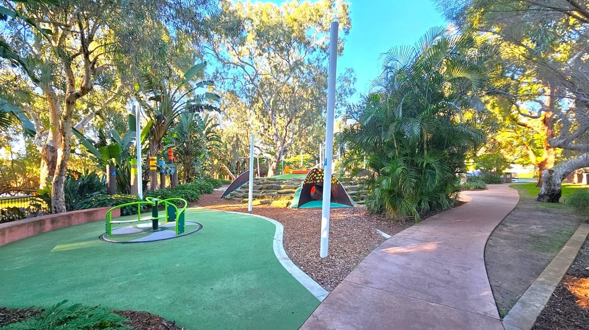 A playground with a green merry-go-round on the left, set on a green rubber surface. In the centre, there's a small hill with a tunnel and climbing features. Tall white poles are scattered around. On the right, a winding concrete path is bordered by lush greenery and trees. The background is filled with more trees, creating a shaded, natural environment.