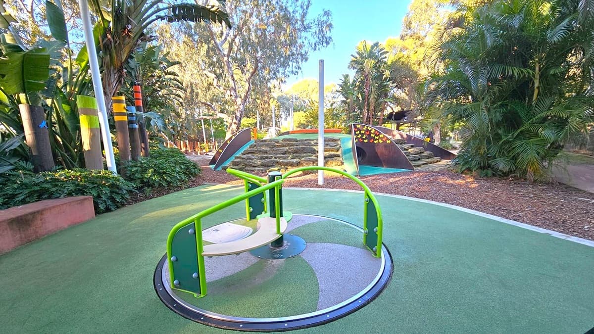 A playground with a green and grey roundabout in the foreground. In the background, there is a mound with a climbing structure featuring colourful butterfly designs. Tall trees and lush greenery surround the area, with wooden posts painted in various colours on the left. The ground is covered with a mix of green safety flooring and mulch.