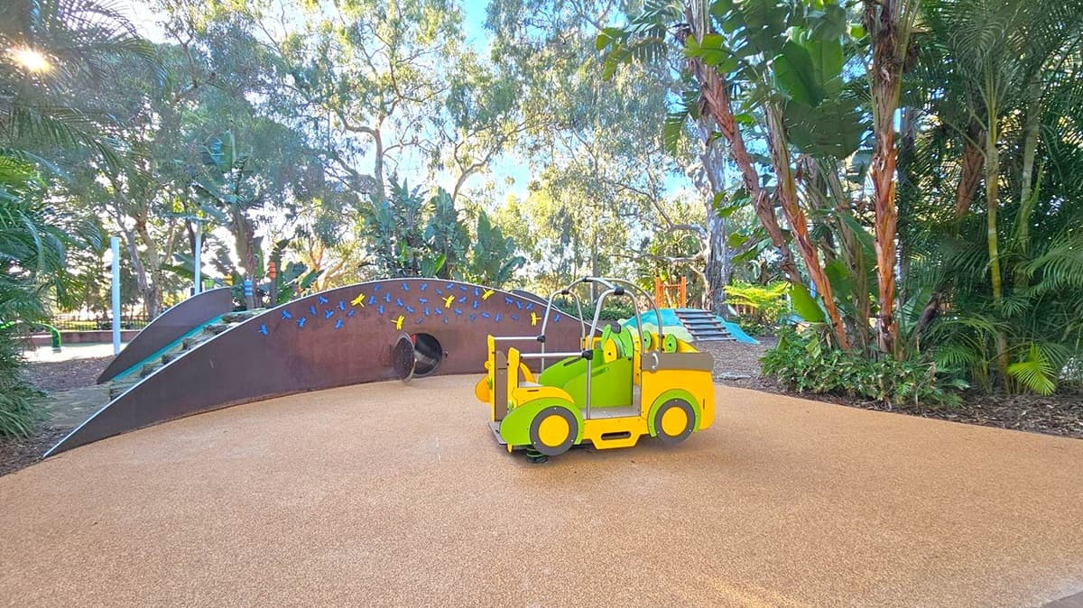 In the foreground, a colourful playground area features a yellow and green toy vehicle on a soft, tan surface. In the middle, a curved climbing structure is adorned with blue and yellow butterfly decorations, with a tunnel entrance visible. To the right, a small set of steps leads up to another play area. The background is filled with lush greenery, including tall trees and dense foliage, creating a natural, shaded environment.