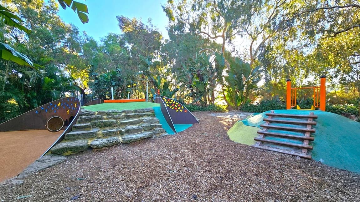 A playground with a natural theme. On the left, there is a slide with colourful butterfly designs and stone steps leading up to it. In the centre, a small hill with a wooden climbing structure is visible. On the right, another hill with a wooden ladder and orange bars at the top. The ground is covered with wood chips, and the area is surrounded by lush green trees and foliage, creating a shaded environment.