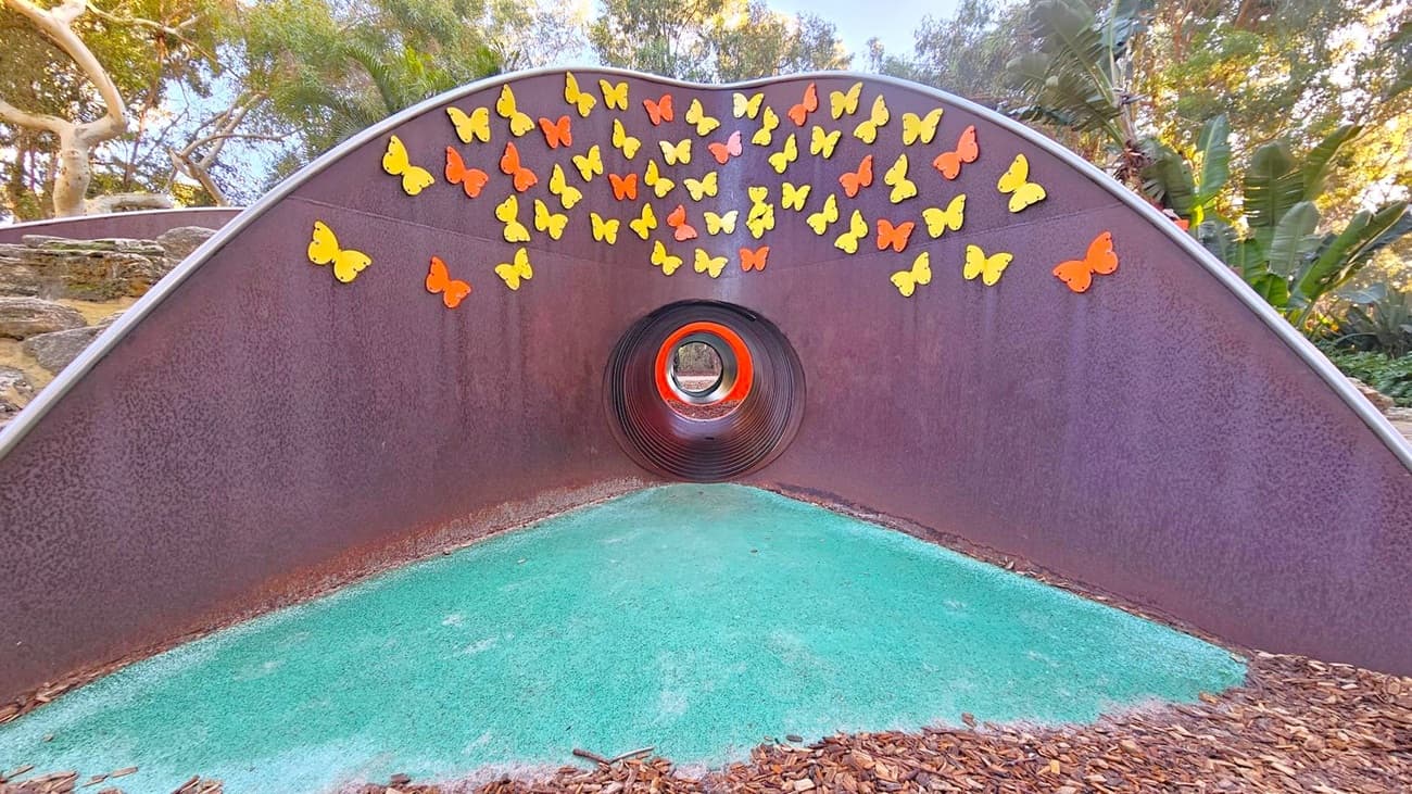 A tunnel structure with a purple exterior and a circular opening in the centre. The wall is adorned with numerous yellow and orange butterfly decorations. The ground is covered with a green surface, and there are wood chips scattered around. In the background, there are trees and foliage, creating a natural setting.
