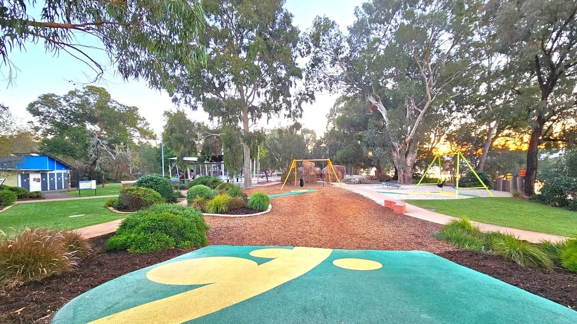 A park with a playground in the centre, featuring swings and a climbing structure. The foreground has a colourful, patterned play surface. On the left, there is a building with a blue roof and public toilets. The right side has a grassy area with another swing set. Tall trees surround the park, and a path winds through the greenery. The sun is setting in the background, casting a warm glow through the trees.