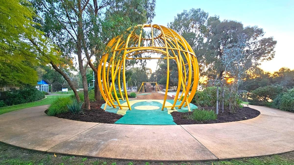A park scene with a large, yellow, spherical metal structure in the centre, resembling a dome. Surrounding it are trees and shrubs, with a paved path curving around the structure. In the background, there is a playground with swings and more trees. The ground is a mix of grass and mulch, and the sky is visible through the trees, suggesting a sunset.