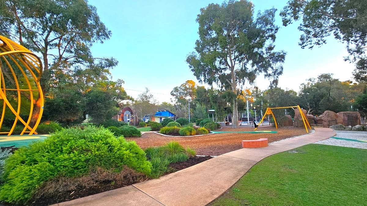 A park with a pathway curving through the centre. On the left, there's a yellow metal structure with curved bars. In the middle, a variety of green shrubs and a large tree are visible. On the right, a yellow swing set stands near some large rocks. The ground is a mix of grass, mulch, and sand. In the background, there are more trees and a building with a blue roof.