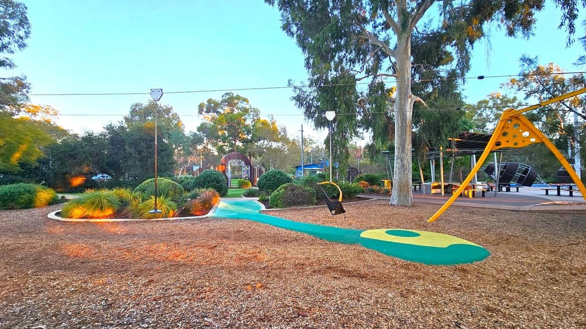 A playground with a zip line in the centre, surrounded by wood chips on the ground. On the right, there's a large yellow climbing structure and a tree. In the background, there are more trees and a pathway leading to a small archway. Benches and a covered seating area are visible to the right. The area is landscaped with bushes and grass, and the sky is clear.