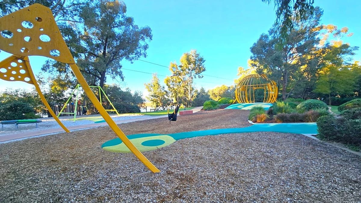 Playground with a large yellow climbing structure on the left featuring circular cutouts. In the centre, a swing set with a black seat is visible. To the right, a spherical yellow climbing frame is surrounded by landscaped greenery. The ground is covered with wood chips and colourful rubber paths. Tall trees and bushes form the background, with a glimpse of water in the distance.