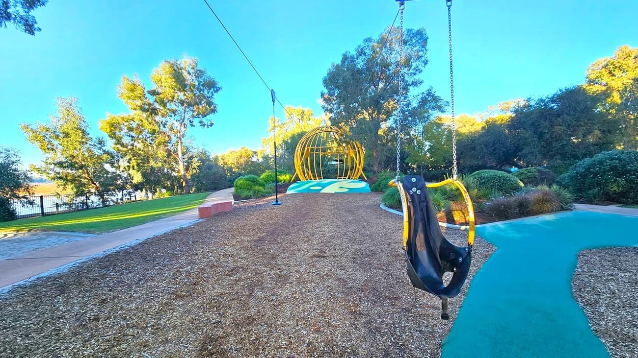 A playground with a zip line on the right, featuring a black and yellow seat suspended from chains. The ground is covered with wood chips, and a path runs alongside. In the background, there's a yellow spherical climbing structure on a green and yellow mound. Trees and bushes surround the area, with a glimpse of a body of water and a fence on the left. The sky is clear and blue.
