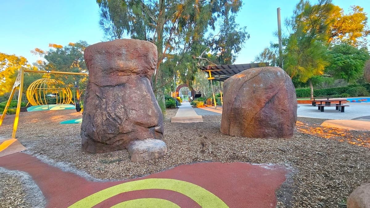 Two large, textured boulders stand prominently in the foreground, with a path running between them. To the left, a yellow metal climbing structure shaped like a dome is visible, set on a colourful play surface. On the right, a picnic table sits on a paved area. Trees with dense foliage surround the scene, and a wooden shelter is partially visible. The ground is covered with wood chips and colourful patterns, and the sky is clear.
