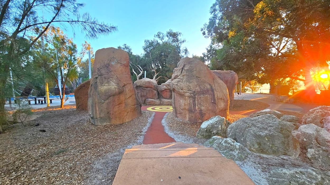 A pathway leads through large, natural rock formations with a circular pattern on the ground in the centre. On the left, there are trees and a playground area with a bench and climbing structures. On the right, more trees are visible, and the sun is setting, casting a warm glow. The ground is covered with mulch and rocks, and the path is bordered by small stones.