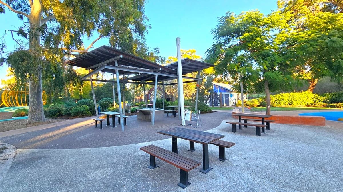 A park setting with several wooden picnic tables and benches on a paved area. In the centre, there is a covered structure with a metal roof supported by poles. To the left, a large tree with hanging branches provides shade. In the background, a spherical yellow climbing frame is visible. On the right, a small building with a blue mural is partially obscured by trees and bushes. The ground is a mix of concrete and gravel, and the area is surrounded by greenery.