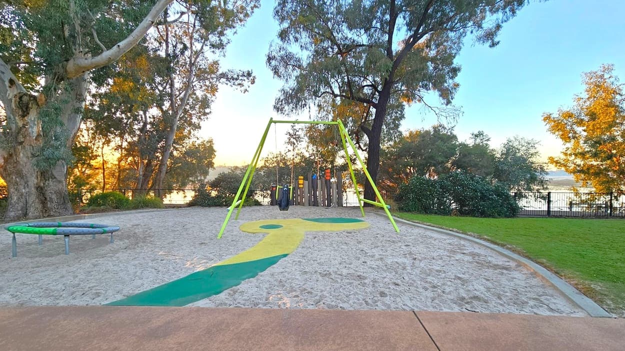 A playground with a sandy area in the centre, featuring a swing set with bright green supports. To the left, there's a circular trampoline-like structure. The background is filled with tall trees and a glimpse of a body of water beyond a fence. The foreground has a paved path leading to the playground, and a patch of green grass is visible on the right. The scene is lit by a warm, low sun, casting long shadows.