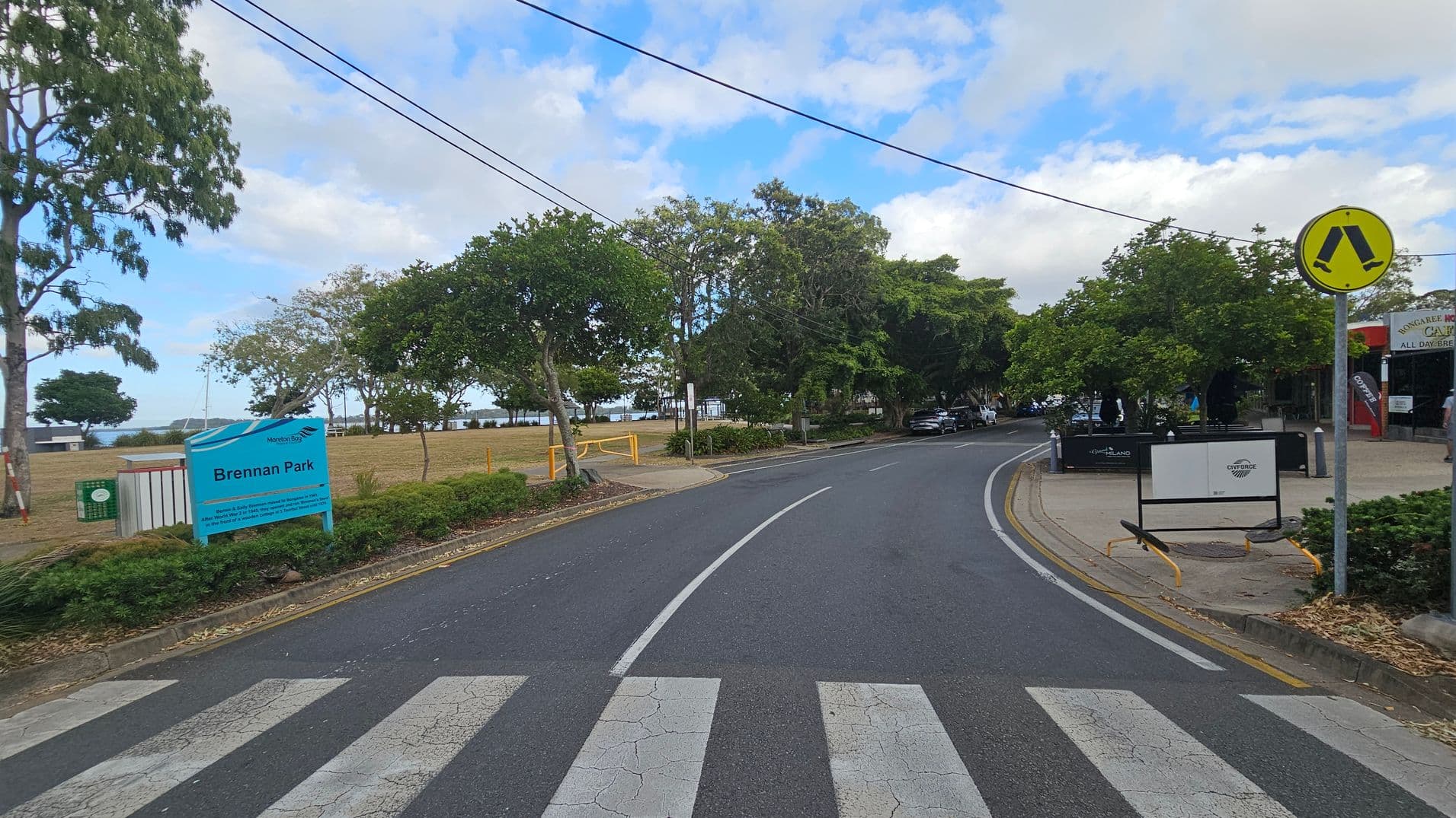 A road curves to the right with a zebra crossing in the foreground. On the left, a sign reads "Brennan Park" next to a grassy area with trees. In the background, there are more trees and a glimpse of water. On the right, a pedestrian crossing sign is visible, along with a footpath and a building with signage. The area is lined with trees and shrubs, and there are parked cars further down the road.