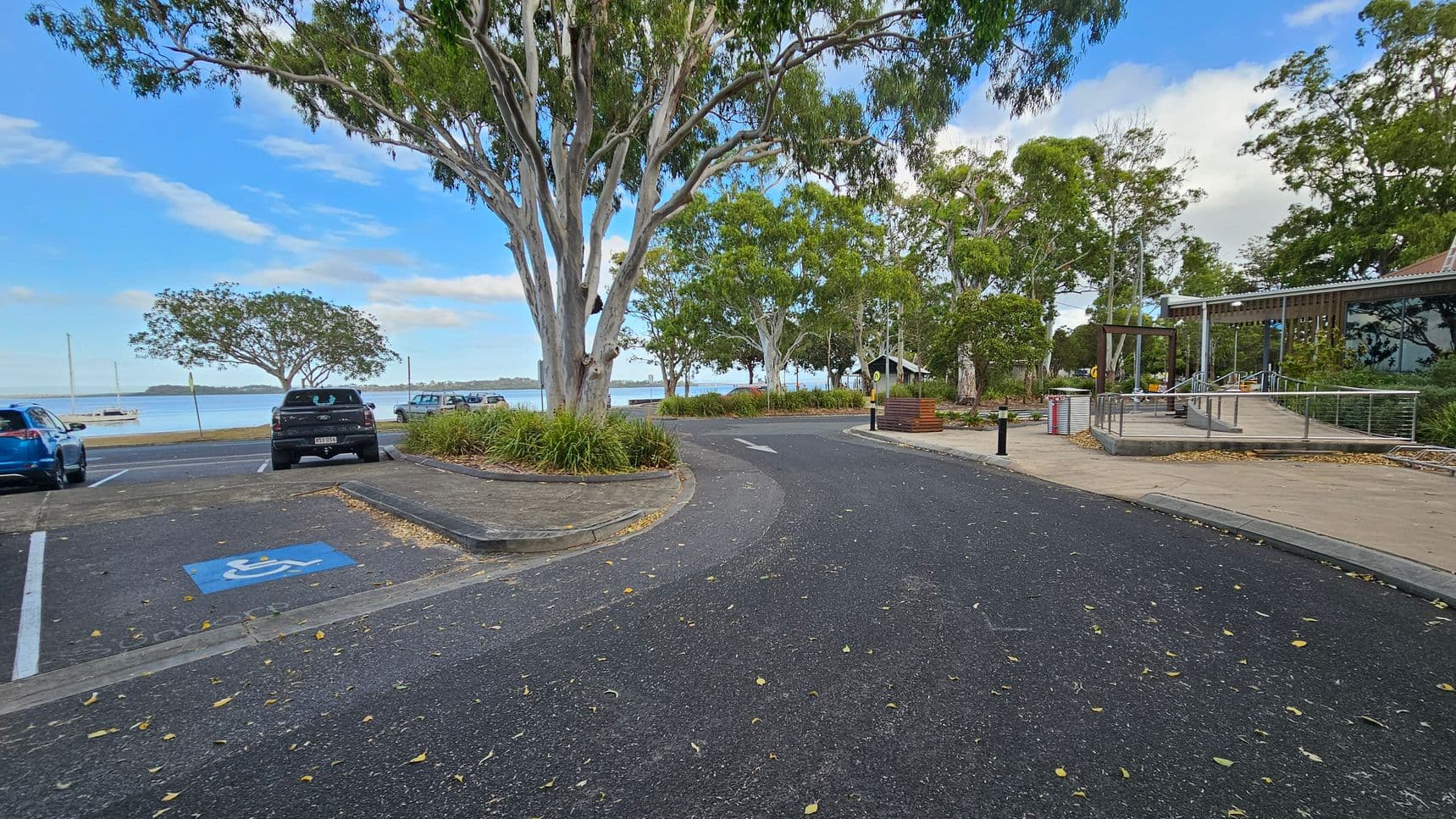 A paved road curves through a park-like area with large trees on both sides. On the left, there are parked cars, including a blue vehicle and a black one in a disabled parking space. In the background, a body of water with a boat is visible. On the right, a modern building with glass windows and a ramp is surrounded by landscaped greenery. The ground is covered with scattered leaves.