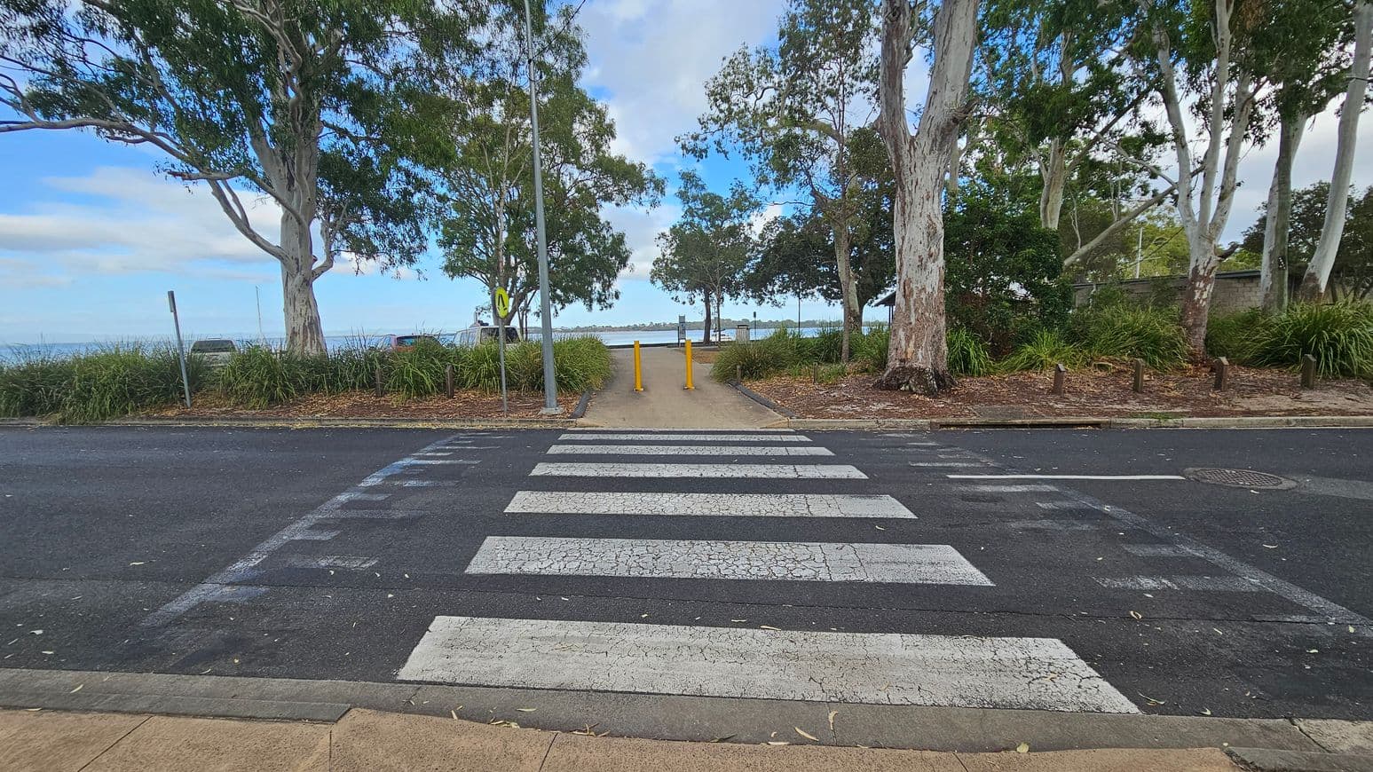 A zebra crossing leads to a path flanked by two yellow bollards. Large trees with thick trunks and green foliage are on both sides, with grass and mulch at their bases. Beyond the path, a body of water is visible in the distance. The road is dark with visible wear, and the pavement is light brown. A metal pole with a sign is on the left side of the path.
