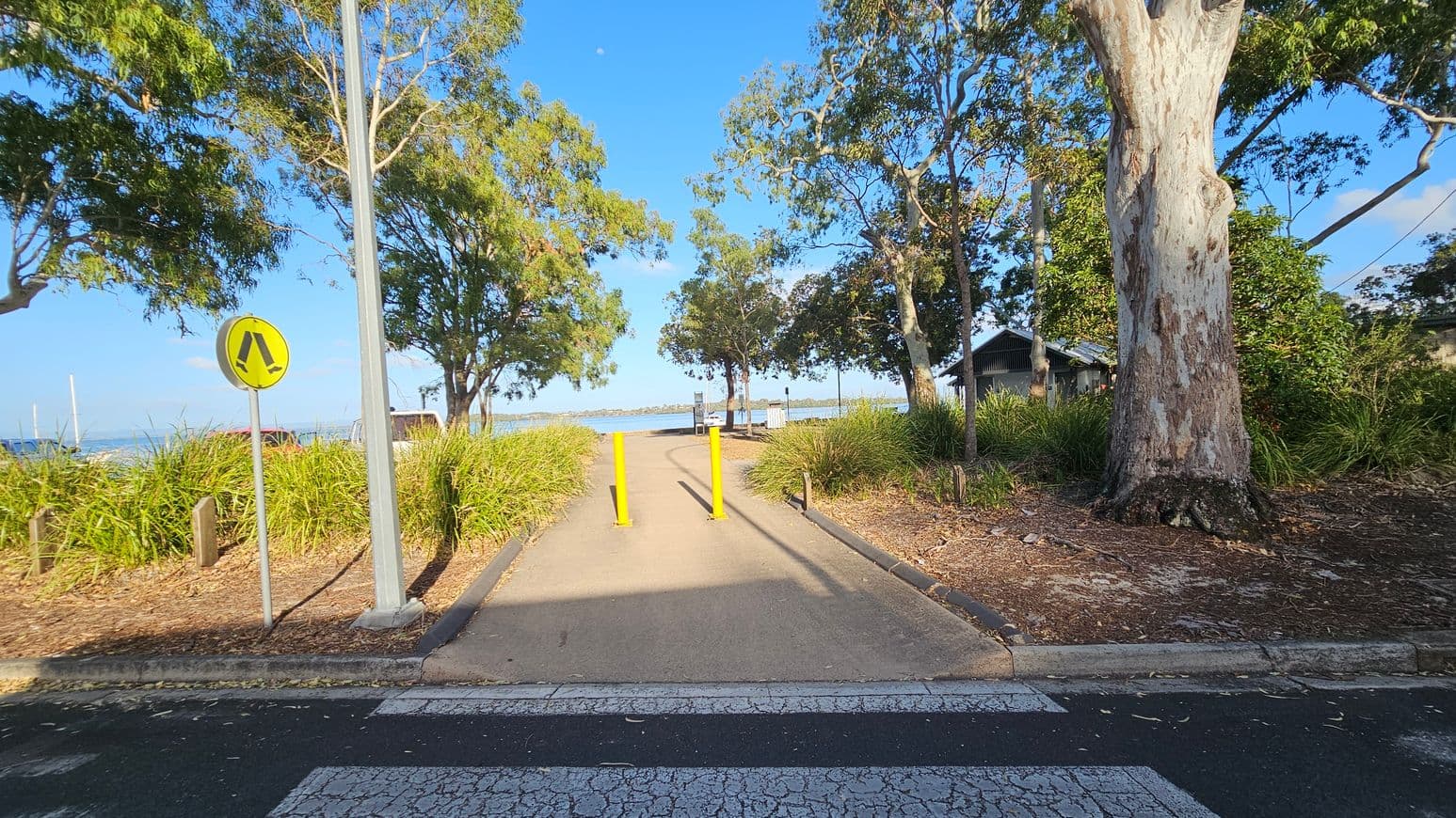 A pathway leads towards a waterfront, flanked by tall trees and dense grass on both sides. A pedestrian crossing sign is on the left, and a large tree trunk is on the right. Yellow bollards are positioned in the middle of the path. In the background, a body of water is visible, with a small structure to the right. The ground is paved, and the area is shaded by the trees.
