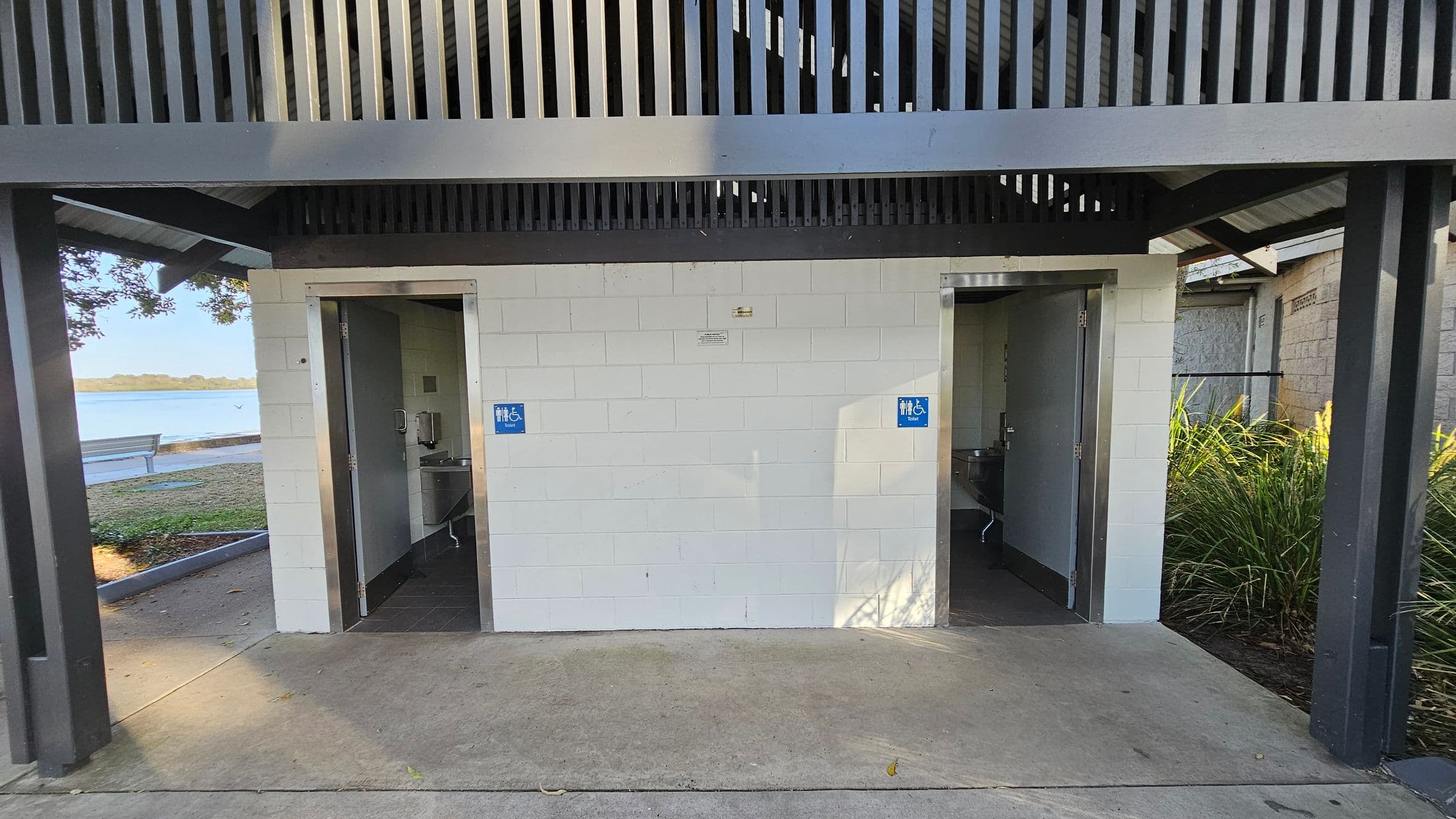 Two public toilets with open doors are visible, each marked with a blue sign indicating accessibility. The structure is made of white bricks with a grey metal roof featuring vertical slats. On the left, a bench and a body of water are visible in the background. On the right, there are green plants. The floor is concrete, and the ceiling is not visible.