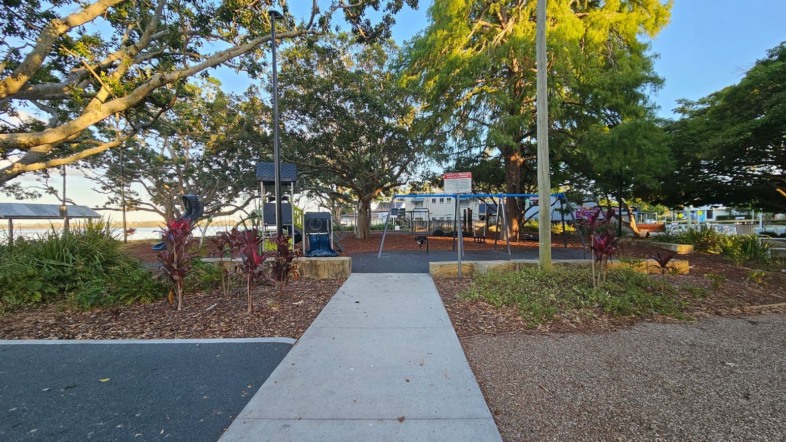 A playground area with a concrete path leading to swings and play equipment. On the left, there are trees with thick branches and a glimpse of a waterfront with a shelter. On the right, more trees and a parking area are visible. The ground is covered with wood chips and small plants, and there are two tall poles near the path. The background features a mix of greenery and buildings.
