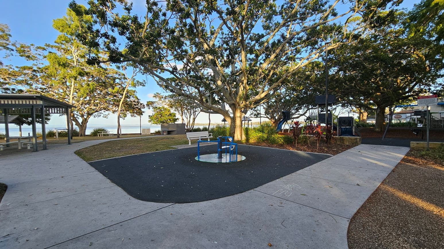 A park scene with a large tree in the centre, surrounded by a paved path. In the middle, there's a blue and white roundabout on a dark rubber surface. To the left, a sheltered picnic area with tables and benches. On the right, a playground with climbing equipment and a slide. The background features more trees and a glimpse of water. The ground is a mix of concrete, grass, and mulch, with scattered leaves.