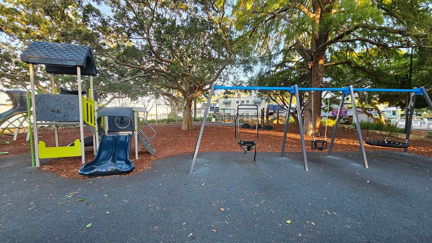 A playground with a play structure on the left featuring a small roof, climbing elements, and a blue slide. To the right, there is a swing set with multiple swings. The ground is covered with a dark rubber surface, and the area is surrounded by trees with dense foliage. In the background, there are buildings and a fence, with some benches and signs visible. The scene is well-lit, suggesting daylight.