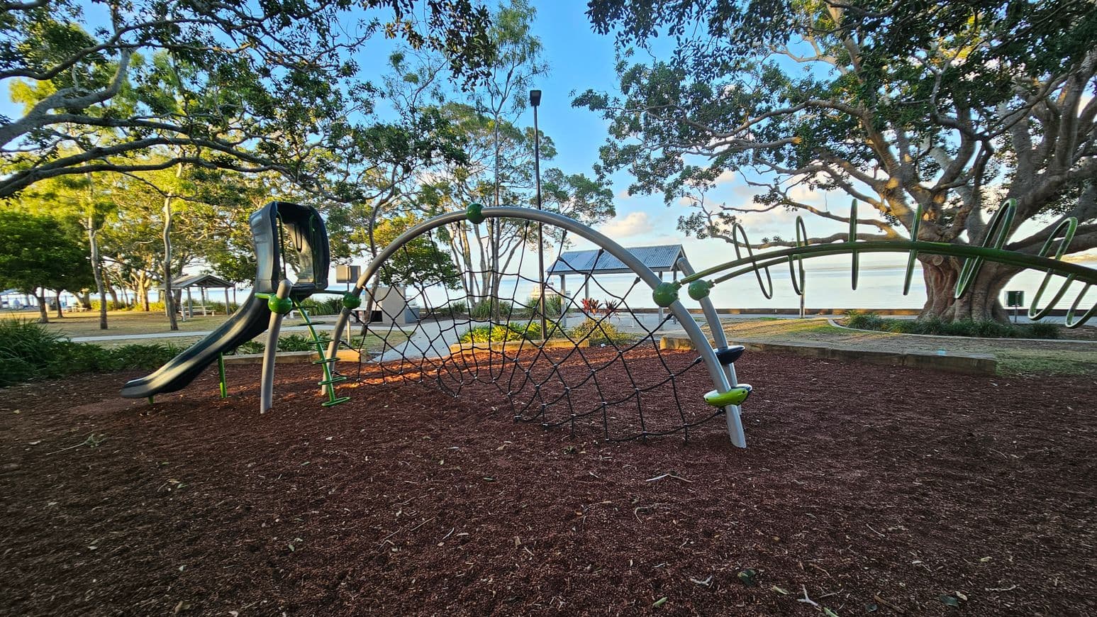 A playground with a slide and climbing net structure on the left, set on a surface covered with reddish-brown mulch. In the background, there are large trees with sprawling branches and a small pavilion with a metal roof. Beyond the trees, a body of water is visible. The area is surrounded by greenery and a paved path runs through the scene.