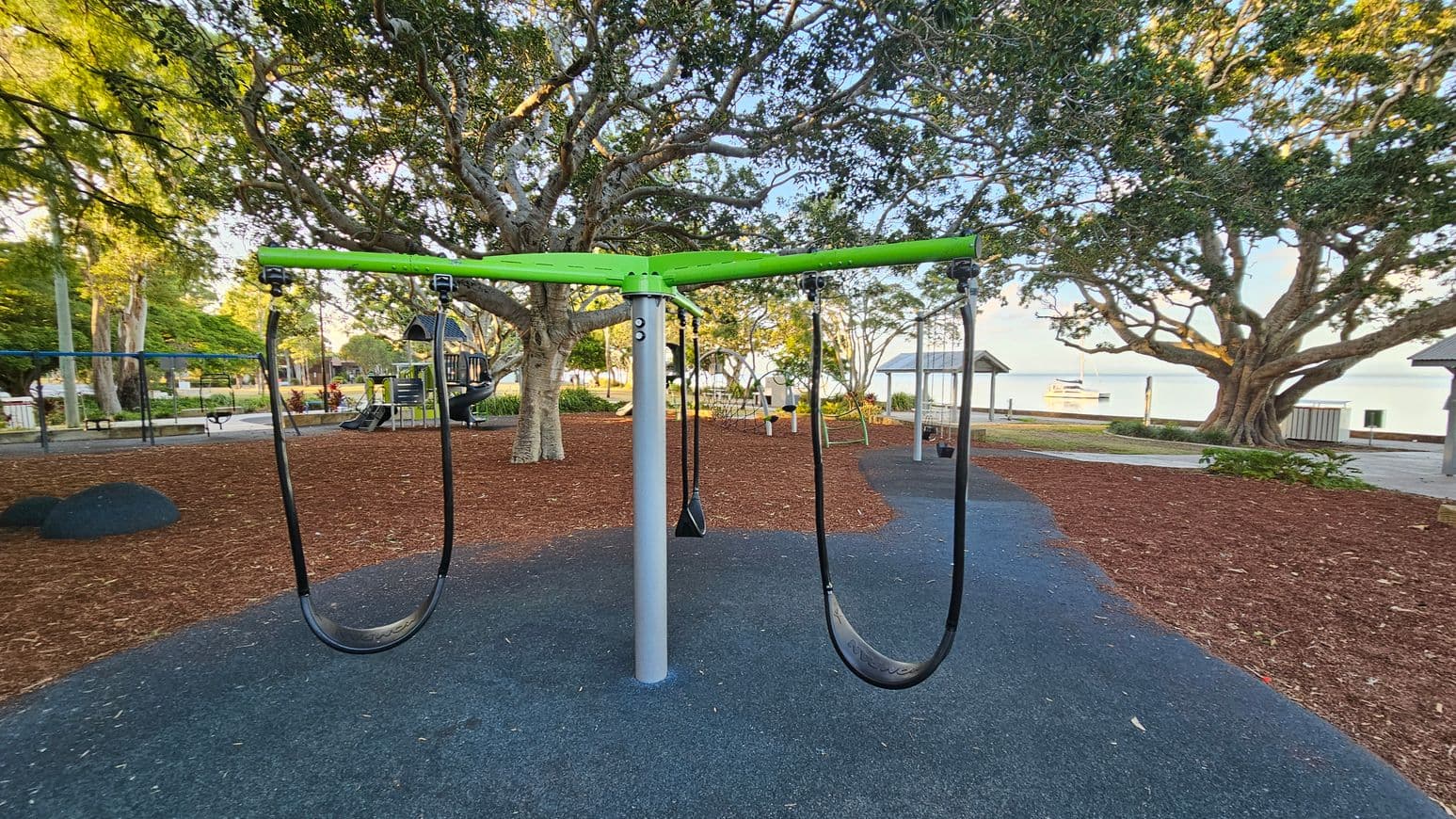 A playground with a central swing set featuring a green top and black seats. The ground is covered with a mix of dark rubber and brown mulch. In the background, large trees provide shade, and a play structure with a slide is visible. To the right, a pavilion and a glimpse of water with a boat can be seen. The area is surrounded by greenery and pathways.