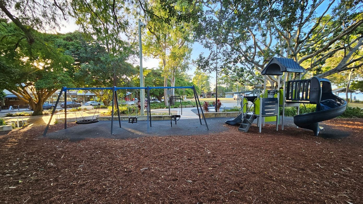 A playground with a swing set on the left, featuring a basket swing and two regular swings. On the right, there's a play structure with a slide and climbing elements. The ground is covered with wood chips, and large trees with dense foliage surround the area. In the background, there are more trees, a pathway, and a few parked cars. The sunlight filters through the leaves, creating dappled light on the ground.