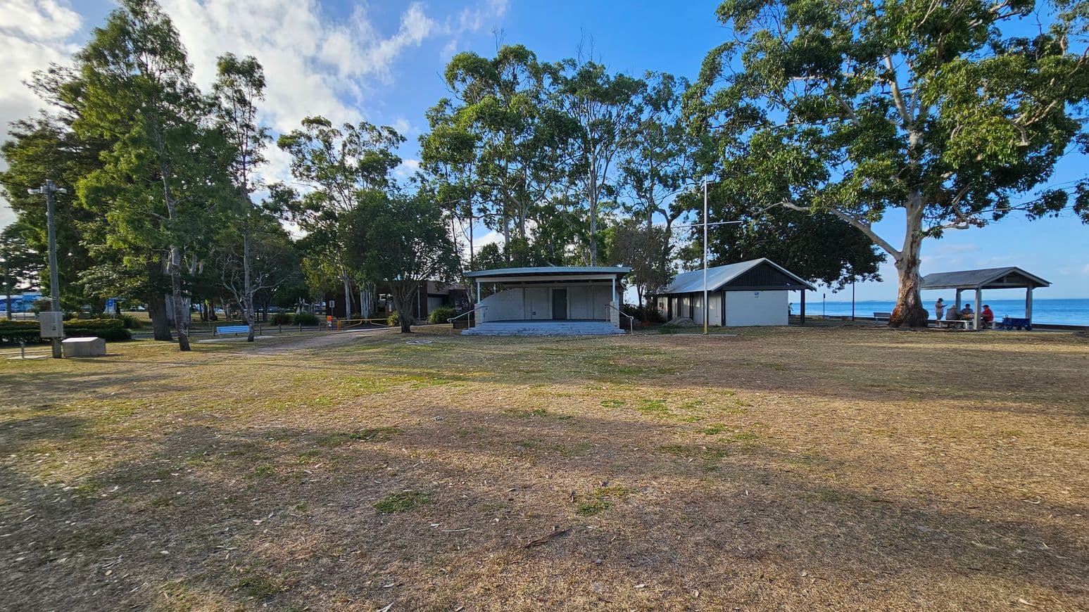 A grassy area with patches of dry soil in the foreground. In the middle, there are two small buildings with metal roofs, surrounded by tall trees. To the right, a group of people sit under a shelter near the water. The background features more trees and a glimpse of the sea. The sky is partly cloudy.