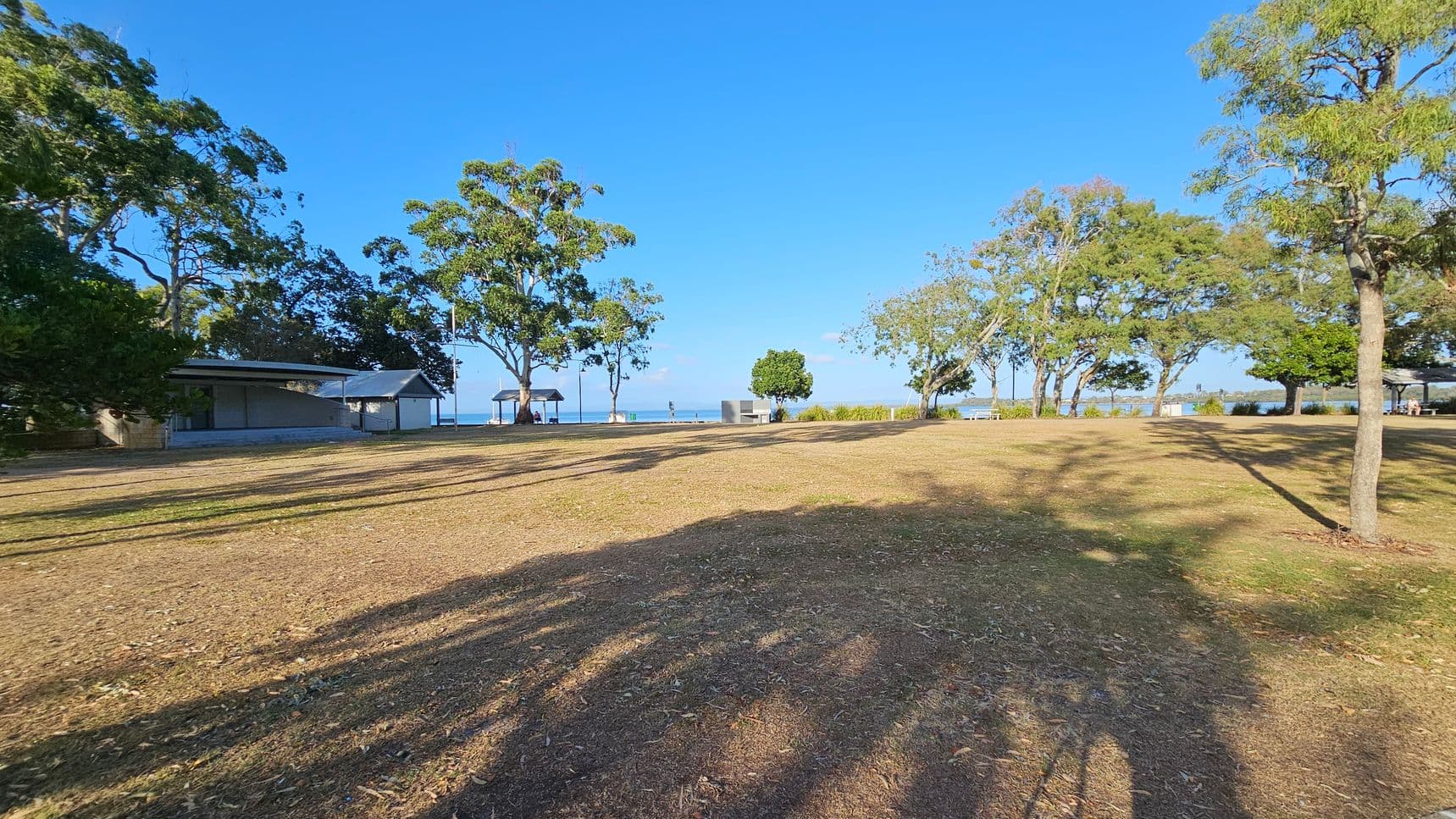 A grassy area with scattered trees casting long shadows. On the left, there is a small building with a sloped roof, partially obscured by trees. In the centre, a path leads towards the water, with a few people visible near a gazebo. On the right, more trees line the edge of the grass, with a glimpse of the sea in the background. The ground is a mix of dry grass and patches of dirt.