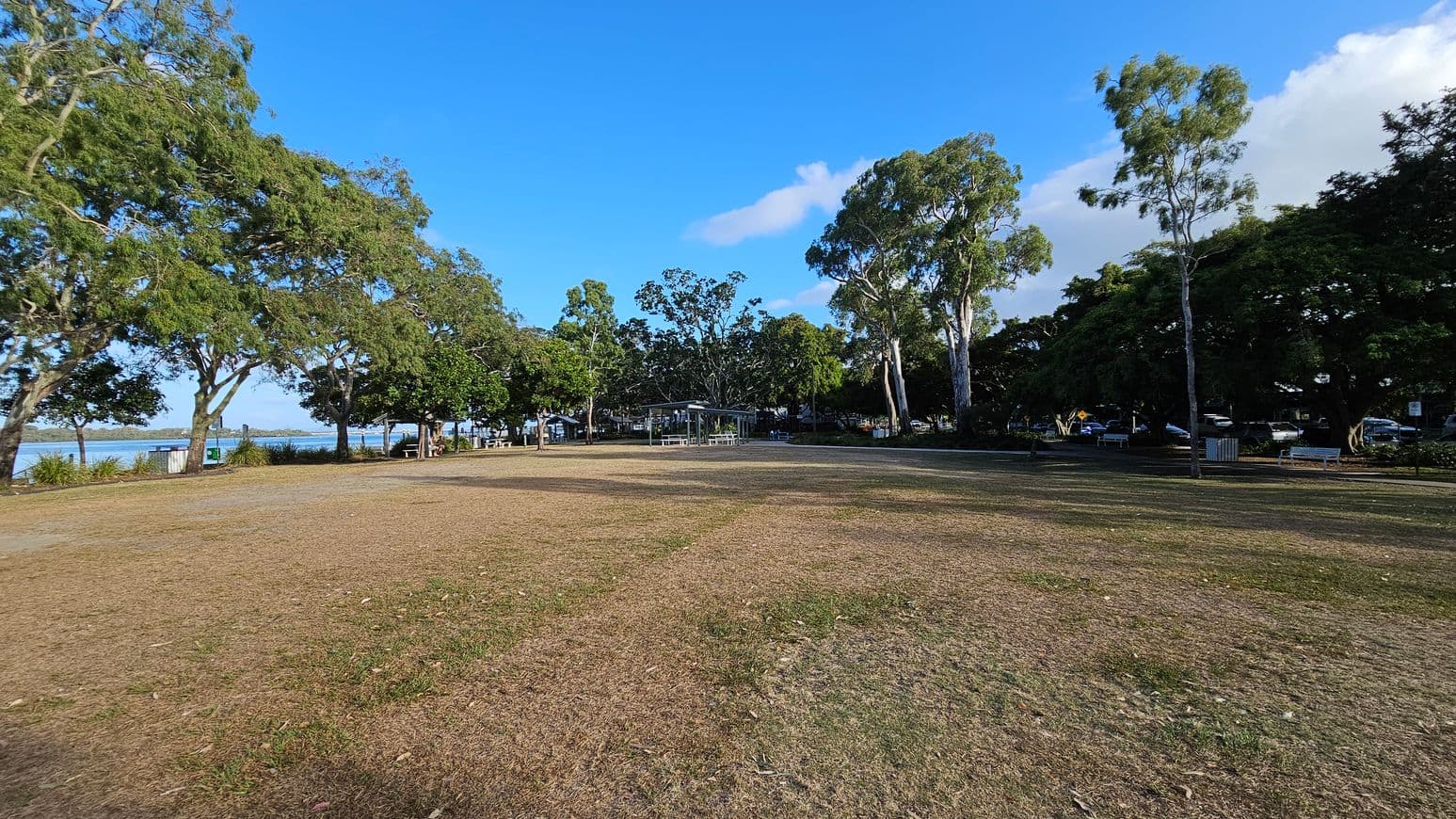 A grassy park area with patches of brown and green grass. On the left, there are large trees with dense foliage, and a glimpse of water is visible in the background. In the centre, a small pavilion with a metal roof is surrounded by more trees. To the right, additional trees and parked cars are visible, along with a few benches and a bin. The sky is clear and blue.