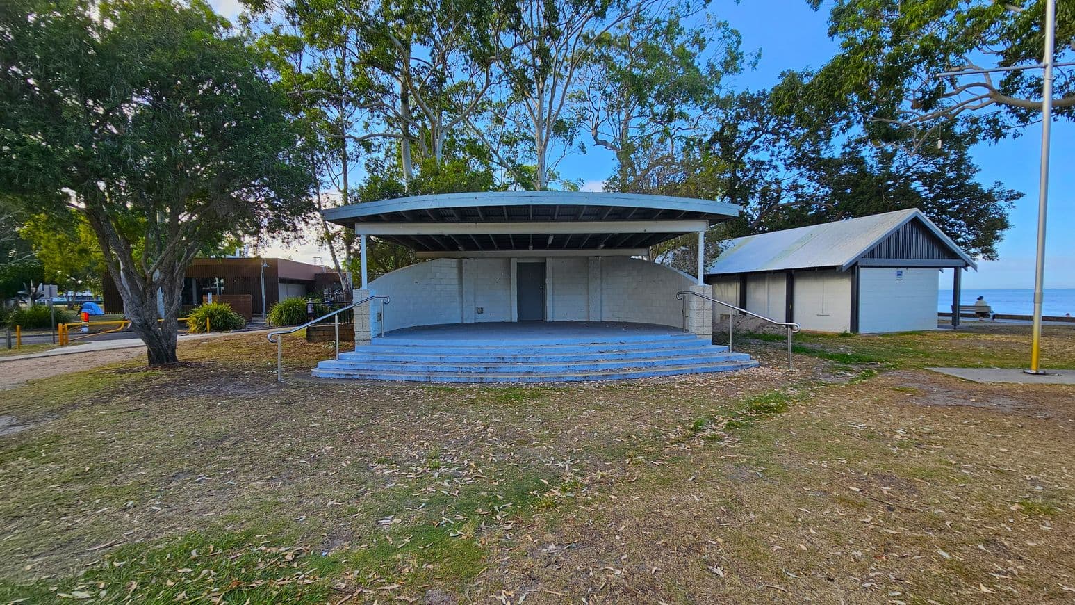 A small outdoor stage with a curved roof and steps leading up to it is in the centre. To the right, there's a small building with a sloped roof. On the left, a large tree provides shade, and a path leads to another structure in the background. The ground is covered with grass and scattered leaves, and a flagpole stands on the right. Trees are visible in the background, and the sea can be seen in the distance.