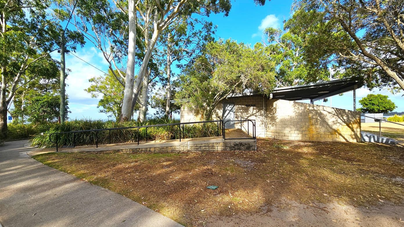 A paved path curves through a park area with tall trees and dense greenery on the left. In the centre, a brick structure with a sloped roof is partially obscured by trees. A metal railing runs alongside a ramp leading to the building. The ground is covered with dry leaves and grass. On the right, a small tree and open grassy area are visible. Shadows from the trees create patterns on the ground and building.