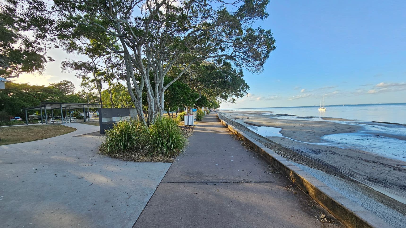 A coastal pathway with trees lining the left side, providing shade over a concrete walkway. To the left, there is a grassy area with a covered seating area and picnic tables. On the right, a low wall separates the path from a sandy beach with a calm sea. A sailboat is visible in the water. The sky is clear, and the area is well-lit, suggesting daytime.