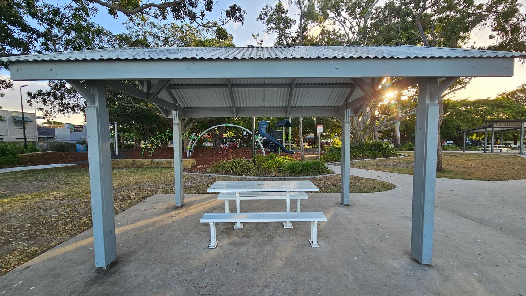A park setting with a metal shelter in the foreground, featuring a corrugated roof and supported by four posts. Underneath is a white picnic table with benches. In the background, a playground with climbing structures and a slide is visible, surrounded by trees and greenery. A path curves around the area, and another shelter is seen to the right. The ground is a mix of concrete and grass, with some mulch around the playground. Sunlight filters through the trees.