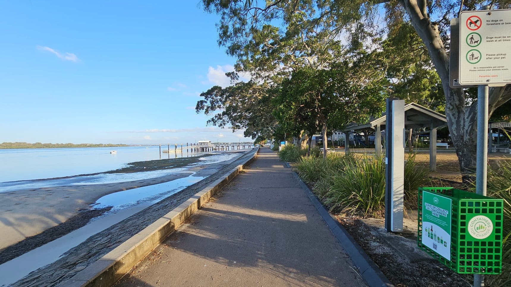 A coastal pathway runs alongside a sandy beach on the left, with a low stone wall separating them. The beach leads to calm water with a distant pier. On the right, a signpost with pet-related rules stands near a green recycling bin. Trees provide shade over the path, and a small shelter with a sloped roof is visible among the greenery. The path is bordered by grass and shrubs, creating a serene, natural setting.