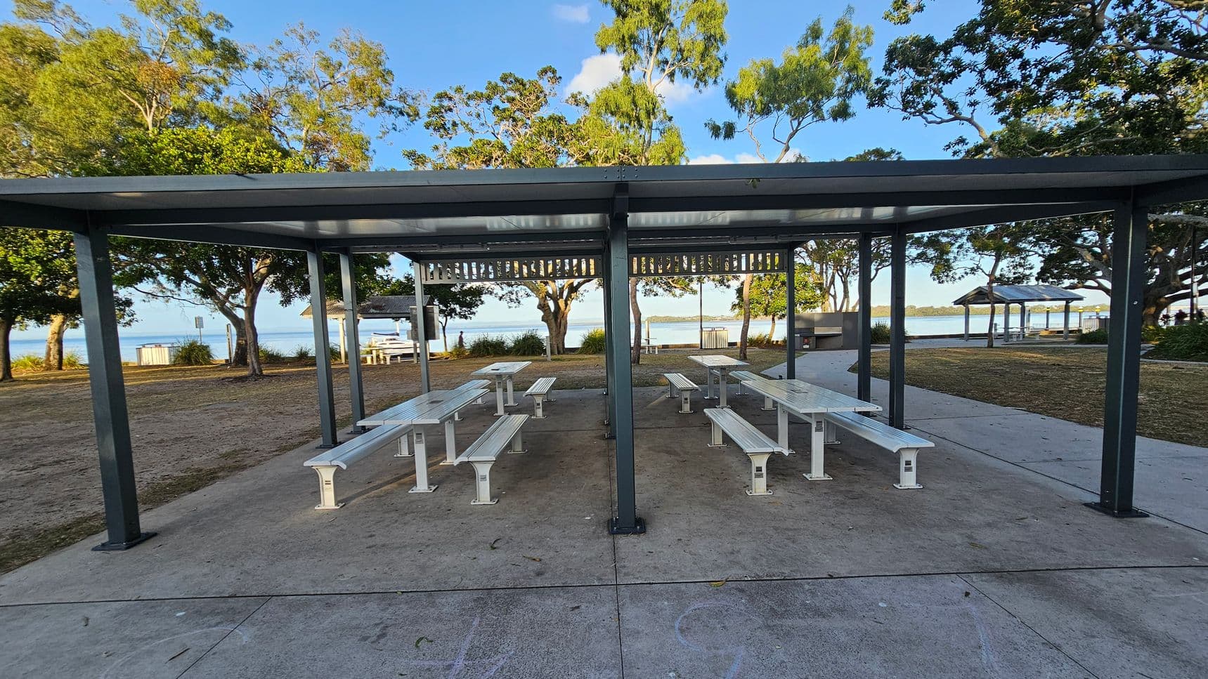 A covered picnic area with several white benches and tables on a concrete surface. The structure has a metal roof supported by dark green posts. In the background, there are trees with green foliage and a view of a body of water. To the right, a pathway leads to another covered area. The ground is a mix of grass and concrete, and the sky is clear.