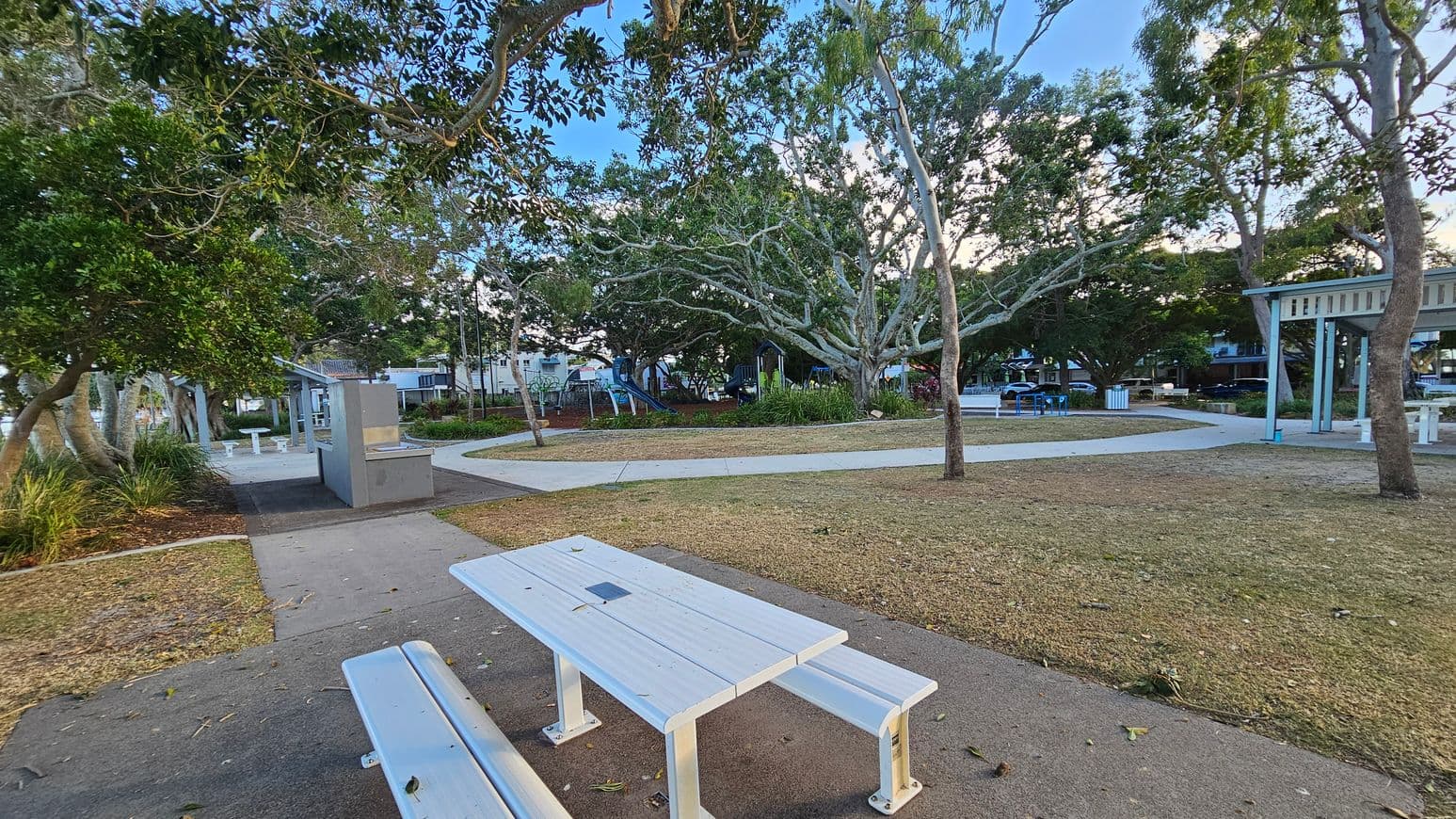 A park with a white picnic table and bench in the foreground on a paved path. To the left, there are trees and a small structure with a barbecue area. In the middle, a playground with a slide is visible among more trees. The background features additional seating areas and a few buildings partially obscured by foliage. The ground is a mix of grass and mulch, and the area is shaded by large trees.