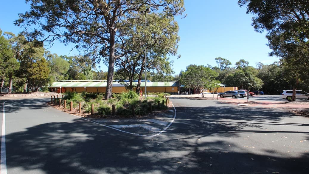 A plain driveway through the Bribie Island Aquatic and Leisure Centre. It is surrounded by beautiful bushland and forest, creating a serene atmosphere. You can spot a street light nearby, as well as two signs that probably look quite alike. The driveway is impeccably maintained, and the broken white lines along the road are clearly visible.