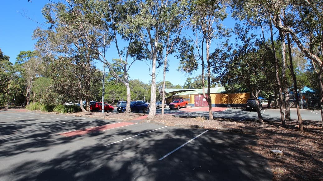 Another angle of the carpark inside the Bribie Island Aquatic and Leisure Centre. The area is filled with tall trees that provide a natural division between the two car spaces. It feels incredibly spacious and well-organized. In the background, you can spot a building painted mostly in yellow, with a striking red front gate.