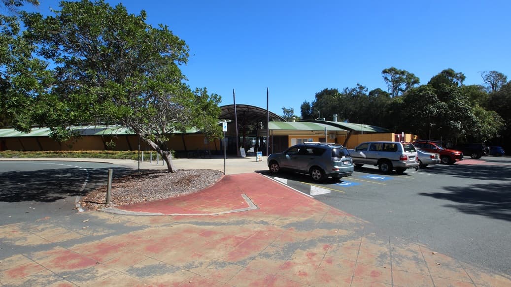 This is the main entrance to the Bribie Island Aquatic and Leisure Centre. In the far right corner, you'll see several parked cars, with two pairs pointing in the same direction. Two poles stand at the very entrance, creating a clear boundary between the outside world and the aquatic and leisure centre beyond.