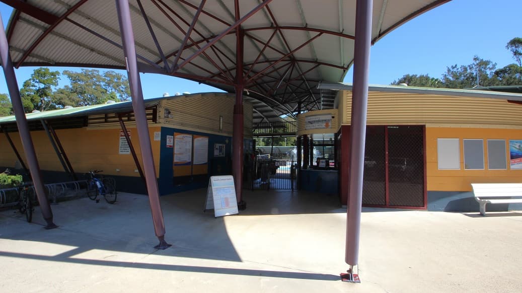 A low-set and long building with curved, corrugated iron roof lines can be seen inside the main entrance. On the left side of the entrance, there is signage and bulletin boards, providing useful information to visitors. As you glance to the right, you'll see a small gate with an entry word prominently displayed at its center. In the left corner of the area, two bikes rest leisurely.