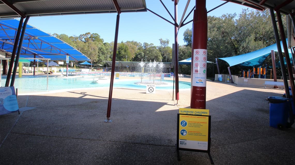 Inside the building, there is a narrow breezeway that serves as a control point for guests entering and exiting. A black pool fence acts as a barrier, indicating that entry requires payment. To the right, there is a low reception counter with a small selection of retail items, such as swimming costumes and goggles. Attached to the reception area is a cozy little café that extends towards the right.