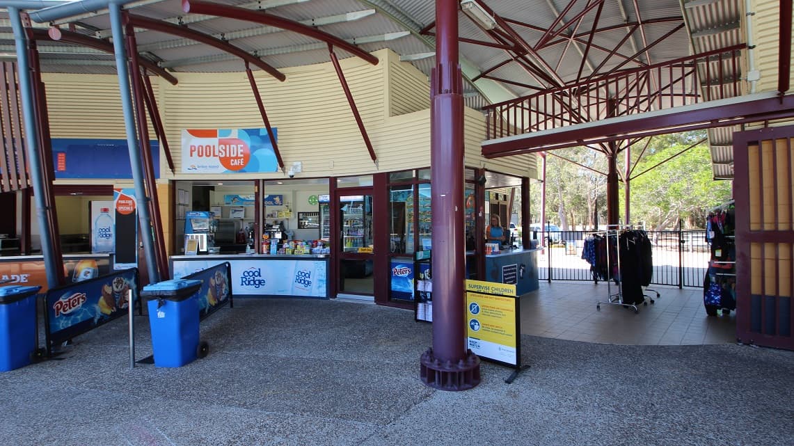 A clearer angle of the reception counter. The low reception counter is to the right and contains a small selection of retail items, including swimming costumes and goggles. A small café is attached to the reception and moves toward the right beyond the reception entry, which is called the poolside café.