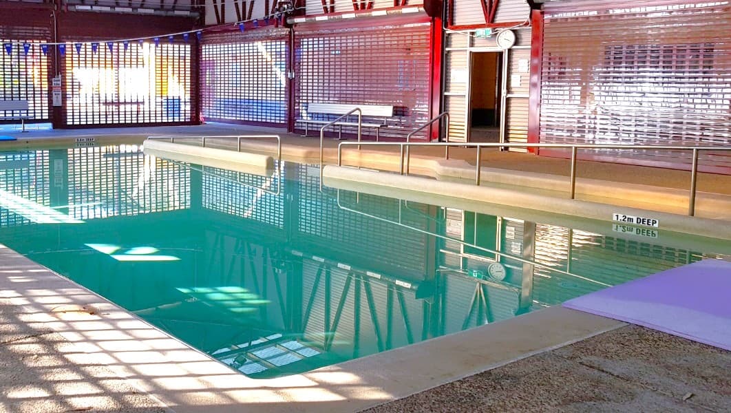 The indoor heated pool at Bribie Island Aquatic and Leisure Centre. There is a small sign indicating that the pool is 1.2m deep. For safety, there are railings placed at the right corner of the pool, right where the main entryway is located. On top of the front door, there's a gallery clock, making it convenient to keep track of time while enjoying the pool.