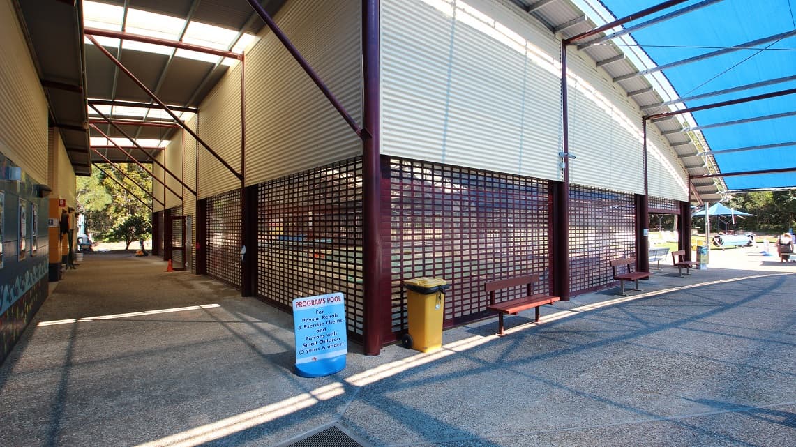 The back angle of the indoor heated pool. Three wooden chairs were placed just outside the pool. There's also a yellow rubbish bin nearby, as well as signage that provides information about pool programs. From this angle, the curved, corrugated iron roof lines are easily noticeable, giving the area a distinct appearance. The back corner of the pool area looks well-kept and tidy.