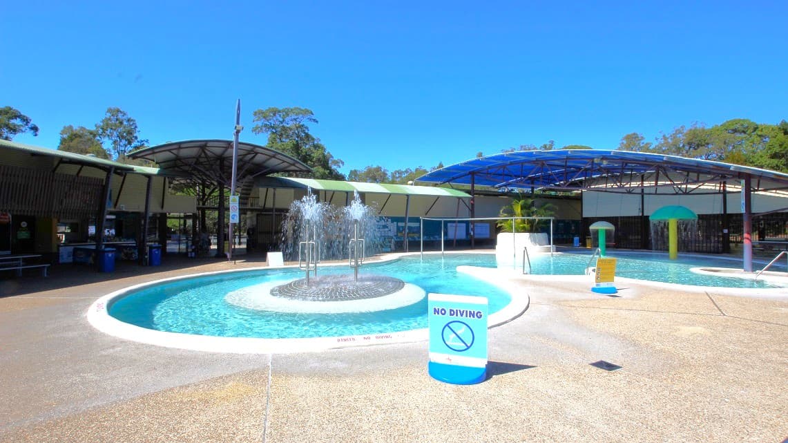 A closer look at the fountain in the open-air aquatic area. It's located right in the center of this pool area, adding a beautiful touch to the surroundings. Just like in other areas, there are signs reminding visitors not to dive. Safety railings can be seen for everyone's safety. On the far right side, there are two mushroom-like decorations, adding a playful element to the pool's atmosphere.