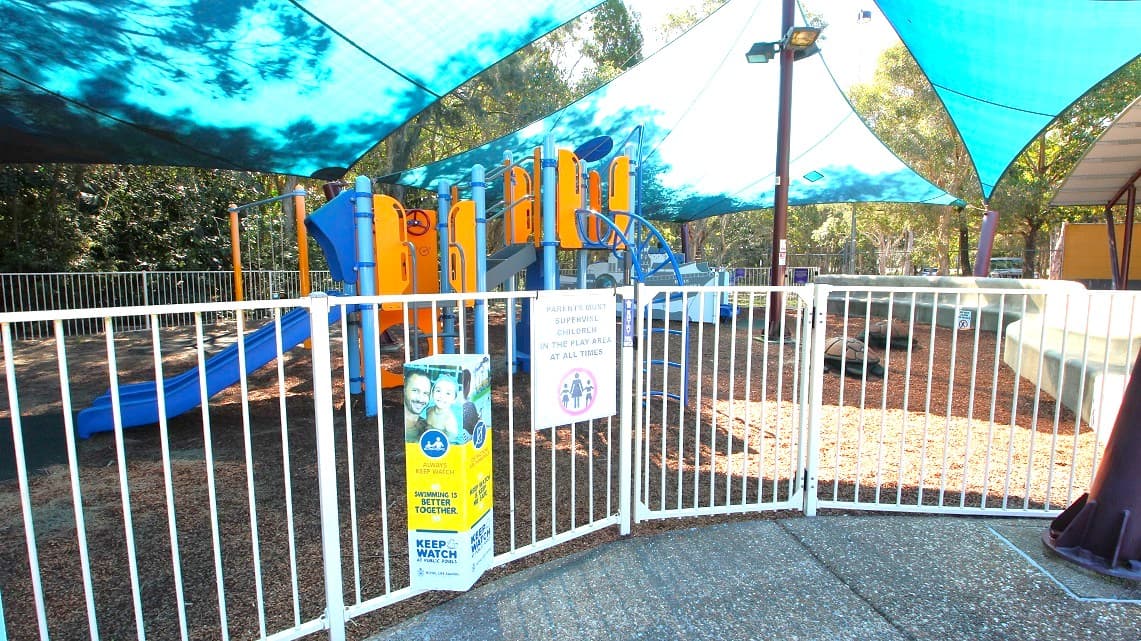 The playground area of the open-air aquatic pool. It's surrounded by trees and covered by white fences. There are signs reminding visitors to keep a close eye on their children. To provide some shade from the sun, three wide pieces of cloth are placed over the area. In the center, there is a post with spotlights, adding some illumination to the space.