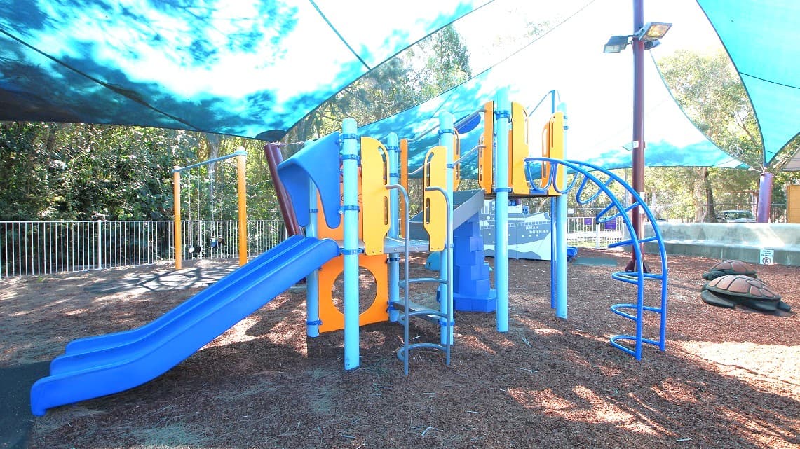 A closer look at the playground area within the pool. It's a spacious space filled with fun activities like slides, swings, and playful decorations such as turtles and a ship. All the playground equipment is brightly colored with shades of blue and orange, adding a vibrant atmosphere. For safety, there are fences surrounding the area, separating it from the thick forests outside.