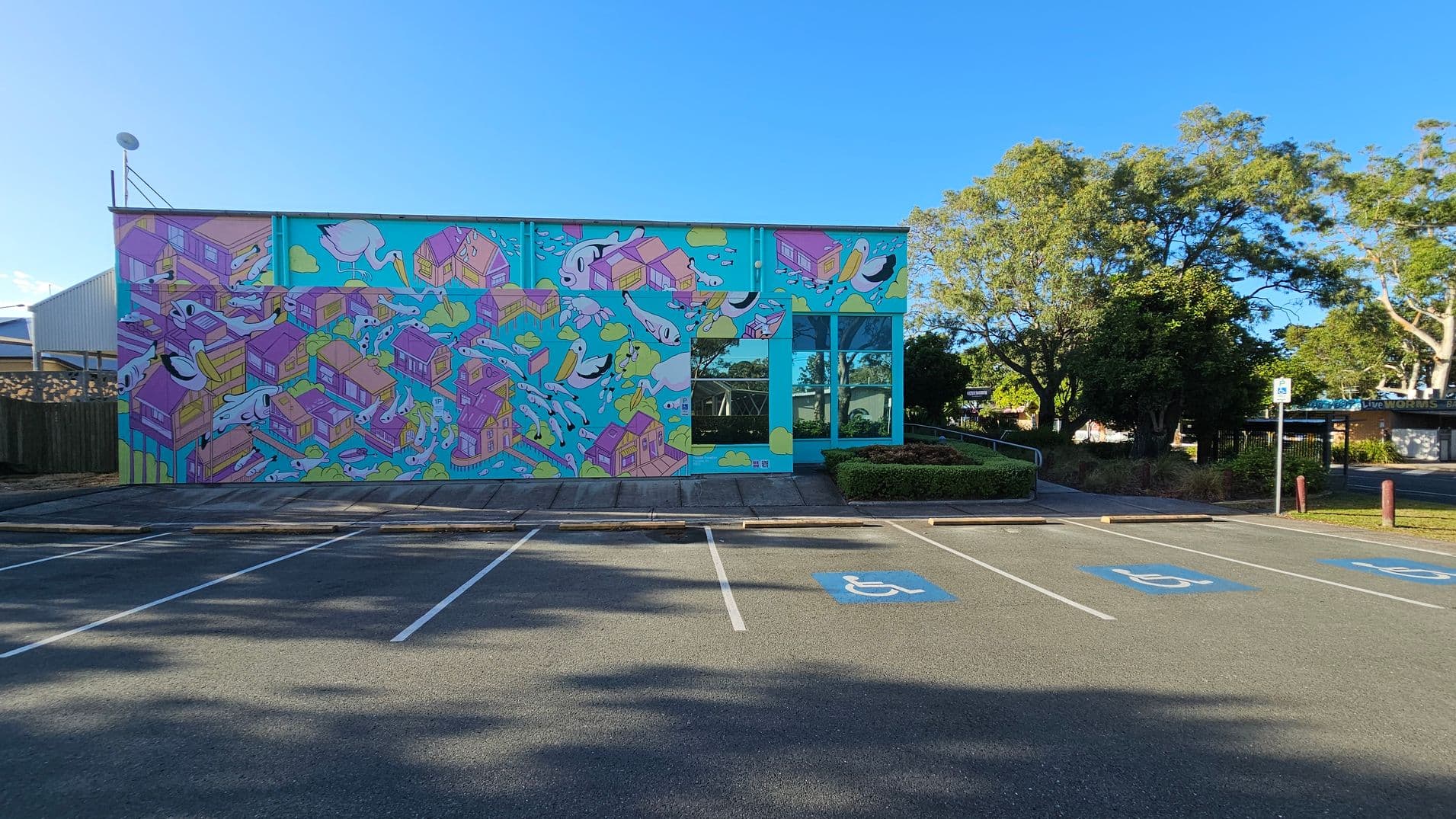 A vibrant mural covers the side of a building, featuring colourful houses and abstract shapes on a bright blue background. To the right, there are large windows reflecting the surrounding greenery. In the foreground, a parking area with marked spaces, including accessible parking spots, is visible. Trees and bushes are in the background, providing a natural setting around the building.