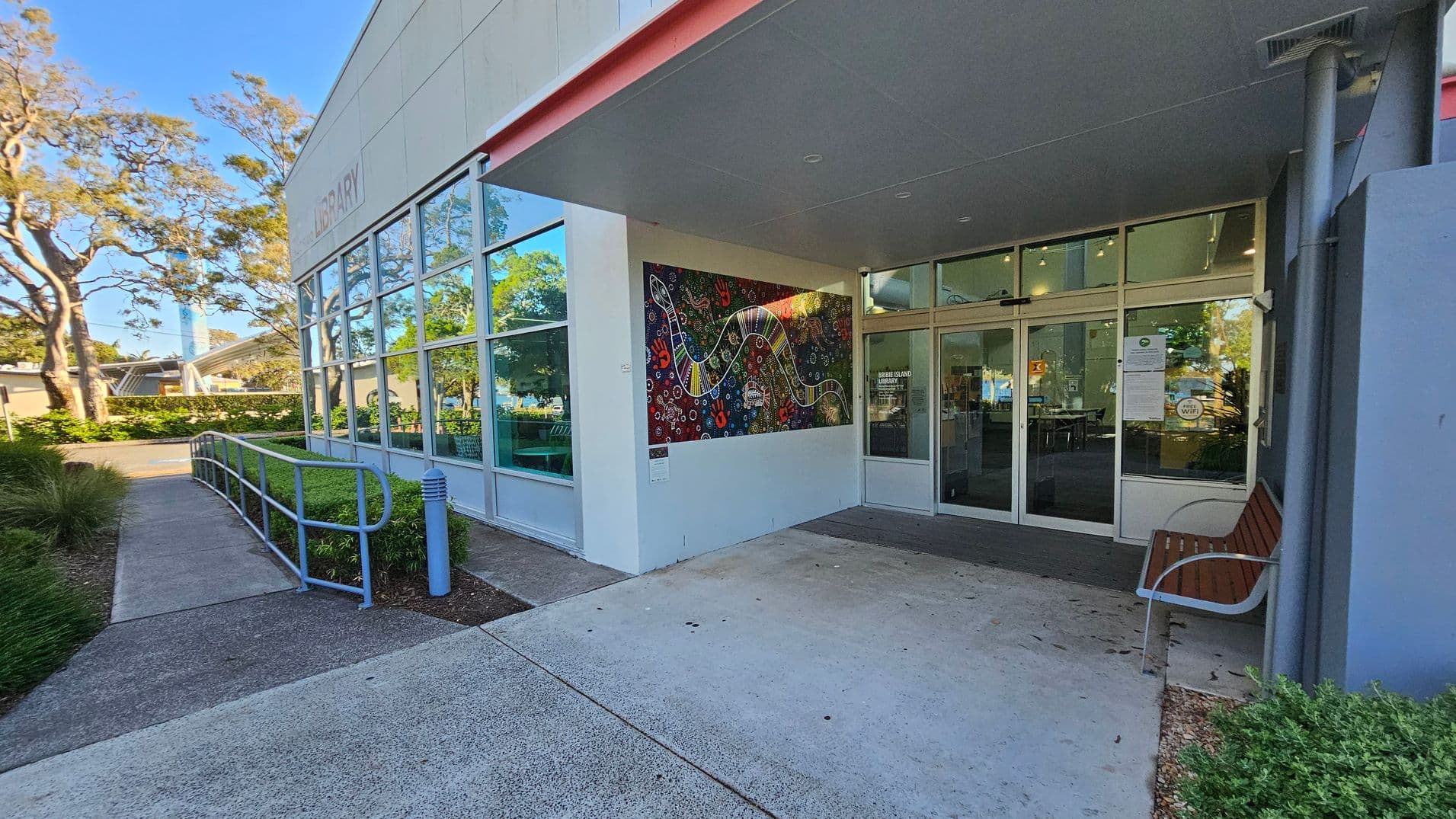 The image shows the entrance to a building with large glass windows and a colourful mural on the wall to the right. A bench is positioned near the entrance, and a pathway with a railing leads up to the door. The building is surrounded by greenery, with trees visible in the background. The ceiling above the entrance is grey, and the ground is paved with concrete.