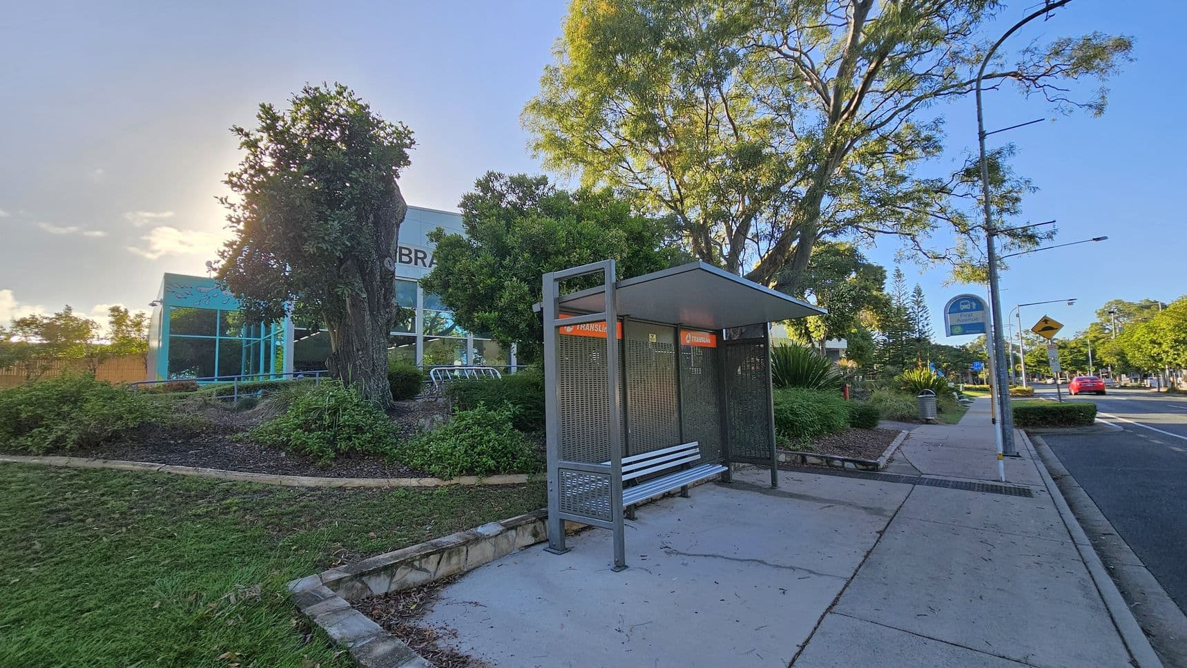 A bus stop with a metal bench and shelter is on the right, next to a paved footpath. A street sign and a rubbish bin are nearby. On the left, a building with large glass windows and the word "LIBRARY" is partially visible, surrounded by trees and landscaped greenery. A large tree stands prominently in the centre. The road runs parallel to the footpath, with a red car in the distance. The sky is clear, and the area is well-lit.